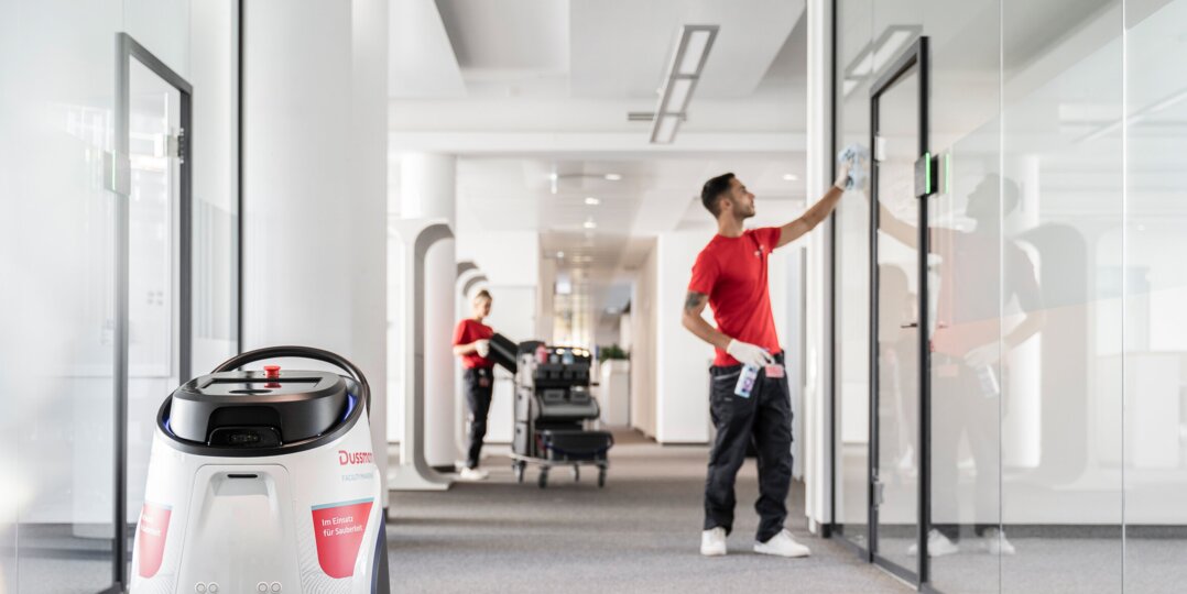 Zwei Reinigungskräfte und ein Reinigungsroboter (Cobotics) in einem Bürogebäude